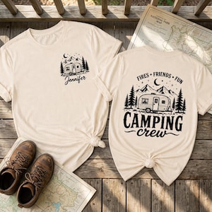 Könnte beinhalten: Zwei cremefarbene T-Shirts mit schwarzen Druckmotiven. Das linke T-Shirt hat einen kleinen Brustdruck mit einem Wohnwagen, Bäumen und dem Namen Jennifer. Das rechte T-Shirt zeigt einen großen Rückendruck mit einem Wohnwagen, Bergen, Bäumen und dem Text Fires, Friends, Fun, Camping Crew.