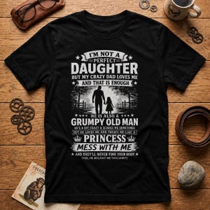 May include: A black short-sleeved t-shirt featuring white text that reads: I'M NOT A PERFECT DAUGHTER BUT MY CRAZY DAD LOVES ME AND THAT IS ENOUGH. HE IS ALSO A GRUMPY OLD MAN HE'S A BIT CRAZY & SCARES ME SOMETIMES BUT HE LOVES ME AND TREATS ME LIKE A PRINCESS MESS WITH ME AND THEY'LL NEVER FIND YOUR BODY (YES, HE BOUGHT ME THIS SHIRT). A graphic of a father and daughter walking is centered.