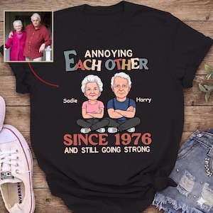 Puede incluir: Camiseta negra con un dibujo animado de una pareja de ancianos, Sadie y Harry, con el texto "Annoying Each Other Since 1976 and Still Going Strong." Una peque&ntilde;a foto de la pareja est&aacute; en la esquina superior izquierda.