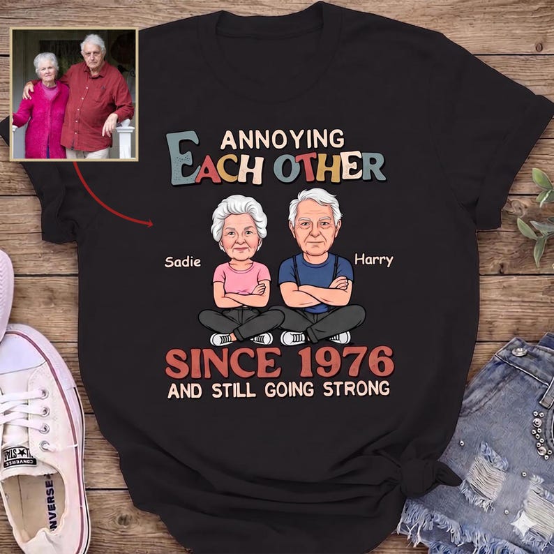 Puede incluir: Camiseta negra con un dibujo animado de una pareja de ancianos, Sadie y Harry, con el texto "Annoying Each Other Since 1976 and Still Going Strong." Una peque&ntilde;a foto de la pareja est&aacute; en la esquina superior izquierda.