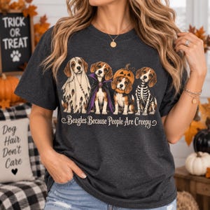 Może przedstawiać: Ciemnoszary melanżowy t-shirt z grafiką przedstawiającą cztery psy rasy beagle w kostiumach na Halloween: ducha, wampira, dynię i szkielet. Pod psami znajduje się napis Beagles Because People Are Creepy wykonany ozdobną czcionką.