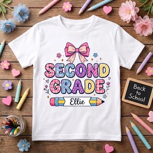 Può includere: Una t-shirt bianca con una grafica colorata che presenta un fiocco rosa sopra il testo "SECOND GRADE" in lettere decorate. Sotto, un disegno di una matita contiene il nome "Ellie". Piccoli fiori e stelle circondano il design sulla maglietta.