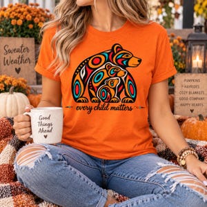 Może przedstawiać: Pomarańczowy t-shirt z krótkim rękawem z grafiką przedstawiającą trzy niedźwiedzie w stylizowanym wzorze sztuki rdzennej w kolorach czerwonym, niebieskim i czarnym. Pod niedźwiedziami znajduje się napis every child matters czcionką kursywą.