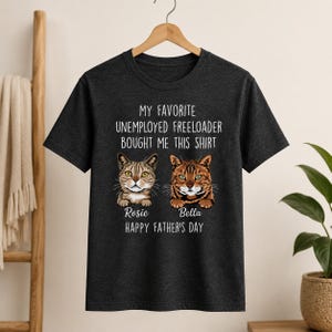 May include: Dark gray t-shirt with the text "MY FAVORITE UNEMPLOYED FREELOADER BOUGHT ME THIS SHIRT." Below the text are two cat portraits, one labeled "Rosie" and the other "Bella." The text "HAPPY FATHER'S DAY" is below the cat portraits.