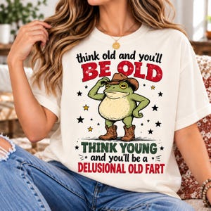 Puede incluir: Una camiseta color crema con el gráfico de una rana verde que lleva un sombrero de vaquero y botas marrones. El texto dice: think old and you'll BE OLD, THINK YOUNG and you'll be a DELUSIONAL OLD FART, rodeado de pequeñas estrellas negras y amarillas.