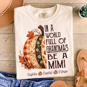 May include: A cream-colored t-shirt featuring a large pumpkin graphic composed of various patterned segments, including plaid and floral prints. The text reads, "IN A WORLD FULL OF GRANDMAS BE A MIMI," with the names "Sophia," "Daniel," and "Ethan" below.