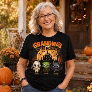 May include: A black short-sleeve t-shirt featuring a graphic of a haunted house, a full moon, and three cartoon monsters: a mummy, a Frankenstein-style creature, and a bat. The text reads "GRANDMA'S LITTLE MONSTERS" with names Daniel, Sophia, and Henry.
