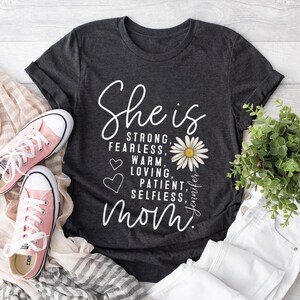 May include: Dark gray t-shirt with the words "She is STRONG, FEARLESS, WARM, LOVING, PATIENT, SELFLESS, Mom" in white script. A daisy graphic and the name "Jennifer" are also featured. Pink sneakers and white jeans are in the background.