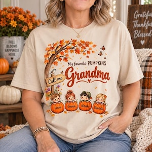 May include: A beige short-sleeve t-shirt featuring a graphic of an autumn tree with orange leaves, butterflies, and a sign reading Call me. Below, four children sit in pumpkins labeled Liam, Emma, Noah, and Charlotte, with text My favorite PUMPKINS Grandma.