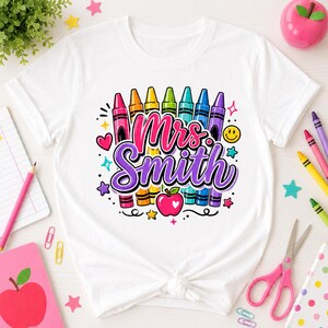 Könnte beinhalten: Ein weißes Kurzarm-T-Shirt mit einem bunten Grafikdruck von sechs Buntstiften in Pink, Orange, Gelb, Grün, Hellblau und Lila. Der Text "Mrs. Smith" ist in einer fetten lila Schreibschrift gedruckt, ergänzt durch ein rotes Apfelsymbol.
