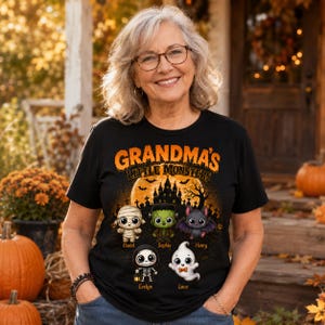May include: A black t-shirt featuring the orange text "GRANDMA'S LITTLE MONSTERS" above a graphic of a haunted house, bats, and five cartoon characters: a mummy, Frankenstein's monster, a bat, a skeleton, and a ghost with names below them.