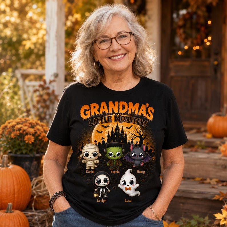 May include: A black t-shirt featuring the orange text "GRANDMA'S LITTLE MONSTERS" above a graphic of a haunted house, bats, and five cartoon characters: a mummy, Frankenstein's monster, a bat, a skeleton, and a ghost with names below them.