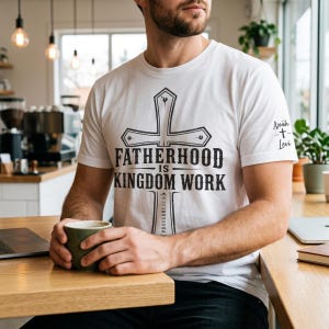 Puede incluir: Camiseta blanca con un diseño de cruz negra y el texto "FATHERHOOD IS KINGDOM WORK". La manga tiene los nombres "Noah + Levi". La persona sostiene una taza verde.
