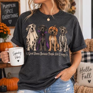 May include: A dark gray t-shirt featuring four Great Dane dogs in Halloween costumes, including a ghost, a vampire, a pumpkin, and a skeleton. Below the dogs, the text reads, Great Danes Because People Are Creepy. A white mug with the text Spooky Vibes is held.