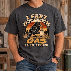 Peut inclure: Un t-shirt gris anthracite à manches courtes avec un motif représentant un corbeau noir tenant une tasse de café. Le texte indique I FART BECAUSE IT'S THE ONLY GAS I CAN AFFORD. Le design inclut un coucher de soleil, des sapins et une branche.