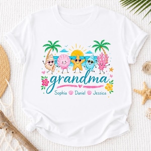 Peut inclure: Un t-shirt blanc à manches courtes avec un motif coloré de créatures marines, de palmiers et d'un soleil. Le mot "grandma" est imprimé en bleu, suivi des prénoms "Sophia", "Daniel" et "Jessica" dans une police bleue plus petite.