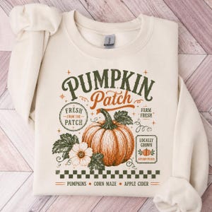 May include: A cream-colored crewneck sweatshirt featuring a graphic print with a large orange pumpkin, green leaves, and text that reads Pumpkin Patch, Fresh From The Patch, Farm Fresh, Locally Grown, Pumpkins, Corn Maze, and Apple Cider.