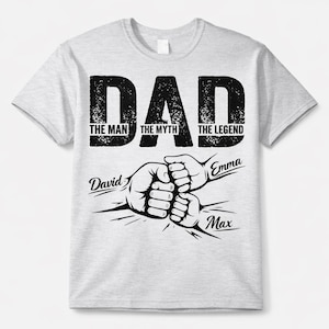 Może przedstawiać: Jasnoszara koszulka z napisem "DAD" dużymi, czarnymi, postarzanymi literami. Poniżej słowa "DAD" znajdują się frazy "THE MAN", "THE MYTH" i "THE LEGEND". Poniżej tekstów znajdują się ilustracje zderzających się pięści, z imionami.