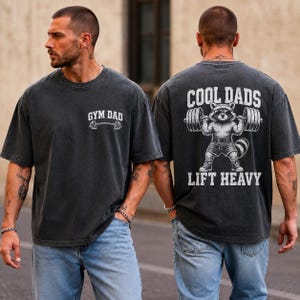 Può includere: T-shirt grigio scuro con la scritta "GYM DAD" e un'immagine di un bilanciere sul davanti. La parte posteriore della maglietta presenta un procione che solleva pesi con la scritta "COOL DADS LIFT HEAVY".