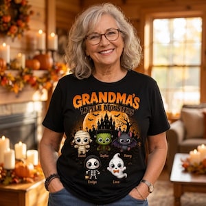 May include: A black t-shirt featuring a graphic print with the text "GRANDMA'S LITTLE MONSTERS" above a haunted house silhouette and five cartoon characters: a mummy, a green monster, a bat, a skeleton, and a ghost, with names below each.