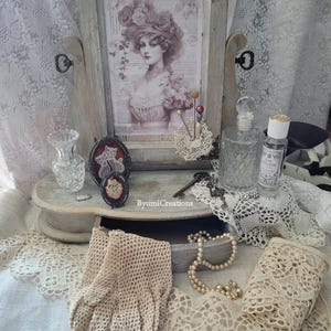 Peut inclure: Un ensemble de toilette de style vintage avec un cadre en bois, un miroir, un tiroir et divers objets décoratifs, dont une estampe encadrée d'une femme, une paire de gants, un collier de perles et des napperons en dentelle.