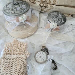 Könnte beinhalten: Ein Stillleben im antiken Stil mit zwei dekorativen Gläsern mit Deckel und floralen Designs, verziert mit Spitze und Bändern. Eine Vintage-Taschenuhr mit Kette und ein Paar gehäkelte Handschuhe sind ebenfalls vorhanden. Die Farbpalette ist weich, mit Creme- und Weißtönen.