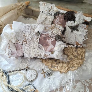 Puede incluir: Dos cajas blancas cubiertas de encaje con fotos antiguas y adornos. Las cajas están sobre un mantel de encaje blanco con un collar de perlas, un reloj de bolsillo y una cruz.