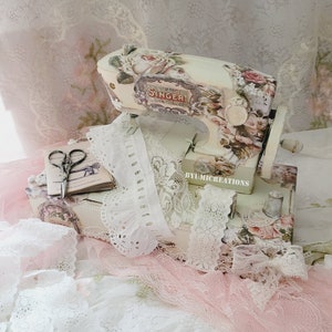 May include: A vintage-style Singer sewing machine, painted white and decorated with floral patterns, sits on a box with lace trim. The box is filled with various types of lace and a pair of scissors.