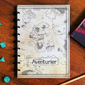 Peut inclure: Un carnet à spirales avec une illustration de carte et le texte "Les archives d'un Aventurier". Plusieurs dés turquoise et un manuel du joueur sont posés sur une surface en bois. Un crayon en bois repose sur la table.