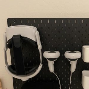 Op de afbeelding: Een zwart pegboard met een witte VR-headset, twee witte controllers, een zwarte case en twee witte opbergbakken. De VR-headset is op het bord gemonteerd, samen met de controllers en de case.