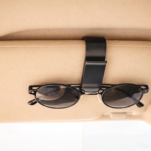 Könnte beinhalten: Schwarze Sonnenbrille, befestigt an einem schwarzen Halter, der an einer beigen Sonnenblende im Auto befestigt ist. Die Sonnenbrille hat einen runden Rahmen und dunkle Gläser. Der Halter ist schlicht und funktional.