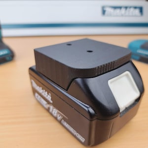 Könnte beinhalten: Ein schwarzer Makita 18V-Akku mit einem schwarzen Adapter. Der Akku hat einen weißen Entriegelungsknopf und weißen Text. Zwei türkis-schwarze Elektrowerkzeuge befinden sich im Hintergrund.