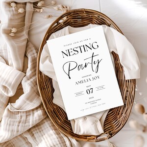 May include: A white card with black text announcing a "Nesting Party" honoring Amelia Joy on Monday, September 7th, 2025 at 6 PM. The card is in a wicker basket with a white onesie.