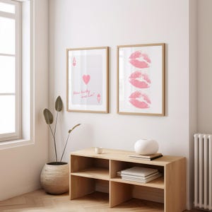 May include: Two framed art prints. One features a playing card design with a pink heart and the text "How lucky are we?". The other print displays three pink lip imprints. Both are in light wood frames.
