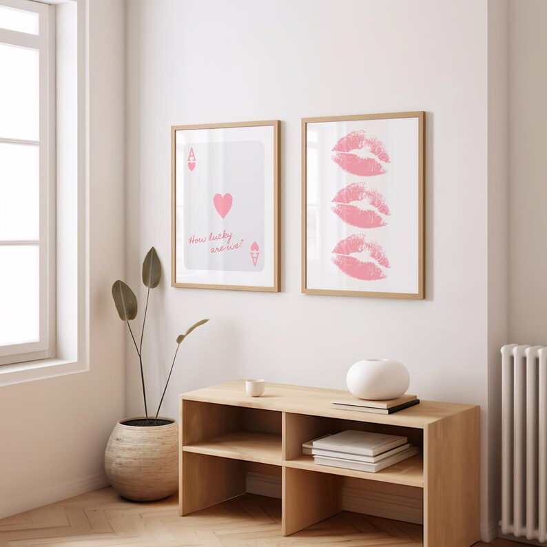 May include: Two framed art prints. One features a playing card design with a pink heart and the text "How lucky are we?". The other print displays three pink lip imprints. Both are in light wood frames.