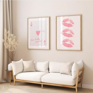 May include: Two framed art prints hang above a white sofa. One print features a playing card design with the text "How lucky are we?" The other print displays three pink lip imprints. The frames are light wood.