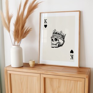 May include: A framed print of a playing card with a black and white illustration of a skull wearing a crown. The card is the King of Spades.