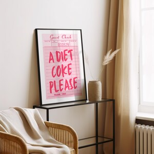 May include: A framed pink print with the words "A Diet Coke Please" in a bold, handwritten style. The print resembles a guest check and is displayed on a black metal table. A beige throw blanket and a wicker chair are in the foreground.