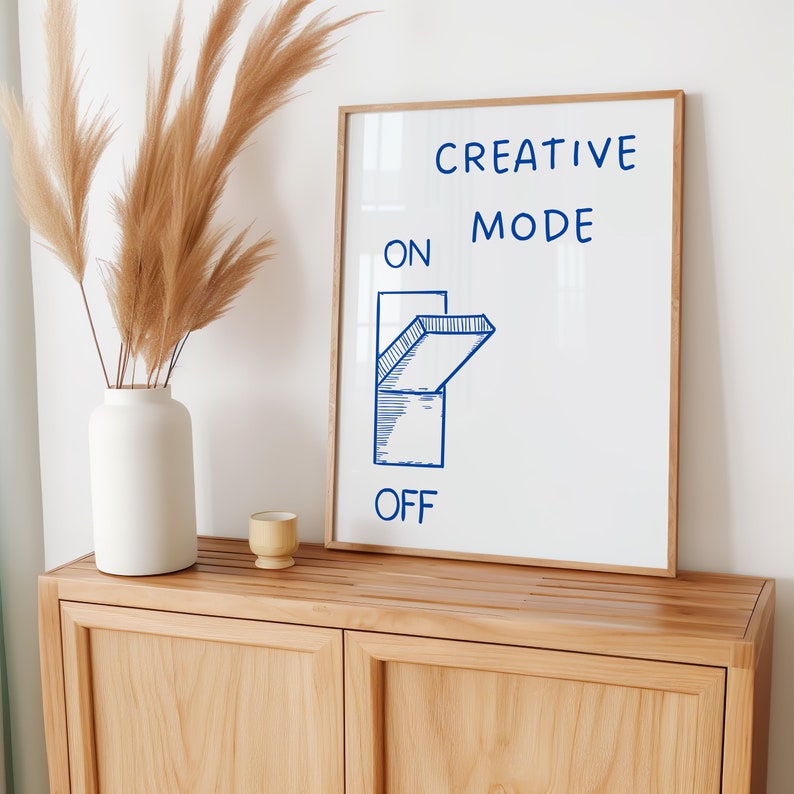 Może przedstawiać: Biały wydruk w ramie z niebieskim tekstem i niebieską ilustracją przełącznika. Tekst brzmi "CREATIVE MODE ON OFF". Przełącznik jest w pozycji "ON". Wydruk leży na jasnym drewnianym szafce.