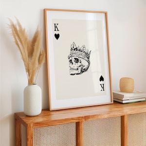 May include: A framed print of a black and white illustration of a skull wearing a crown. The skull is the King of Spades playing card. The card is on a cream background.