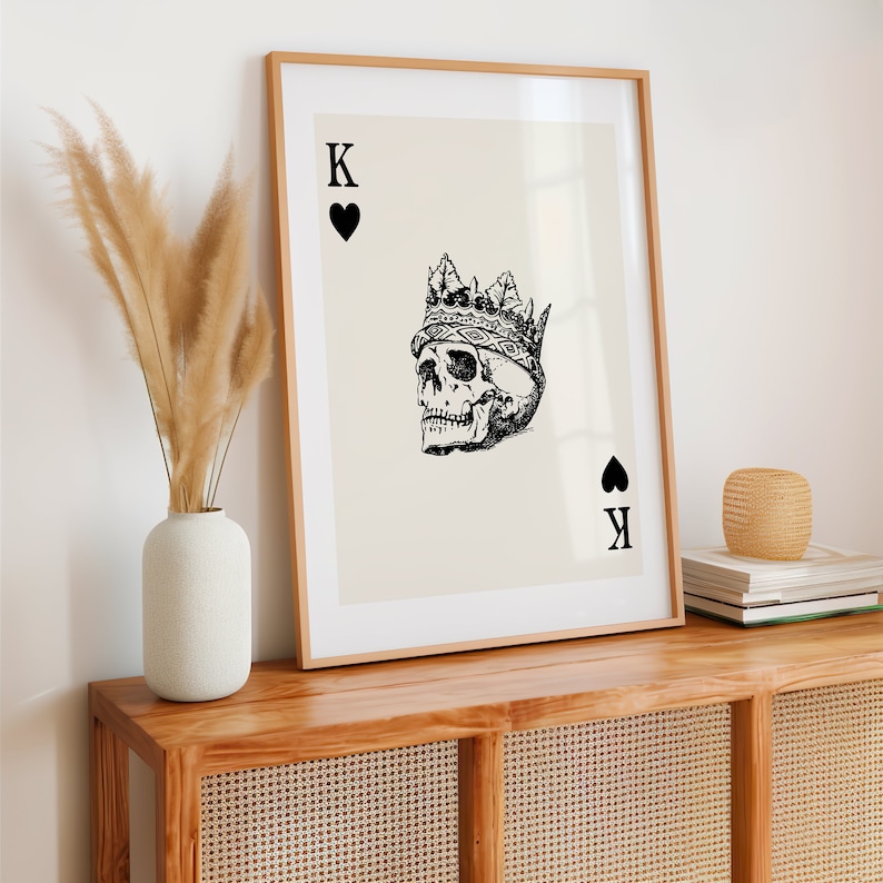 May include: A framed print of a black and white illustration of a skull wearing a crown. The skull is the King of Spades playing card. The card is on a cream background.