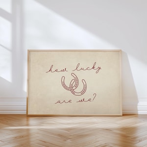 May include: Framed print with the text "how lucky are we?" in a handwritten style. Two red horseshoes are centered below the text. The print has a beige background and a wooden frame.