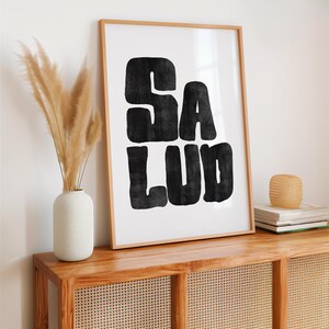 Op de afbeelding: Een zwart-wit print met het woord "SALUD" in een retro, vet lettertype. De print is ingelijst in een licht houten lijst en hangt aan een witte muur.