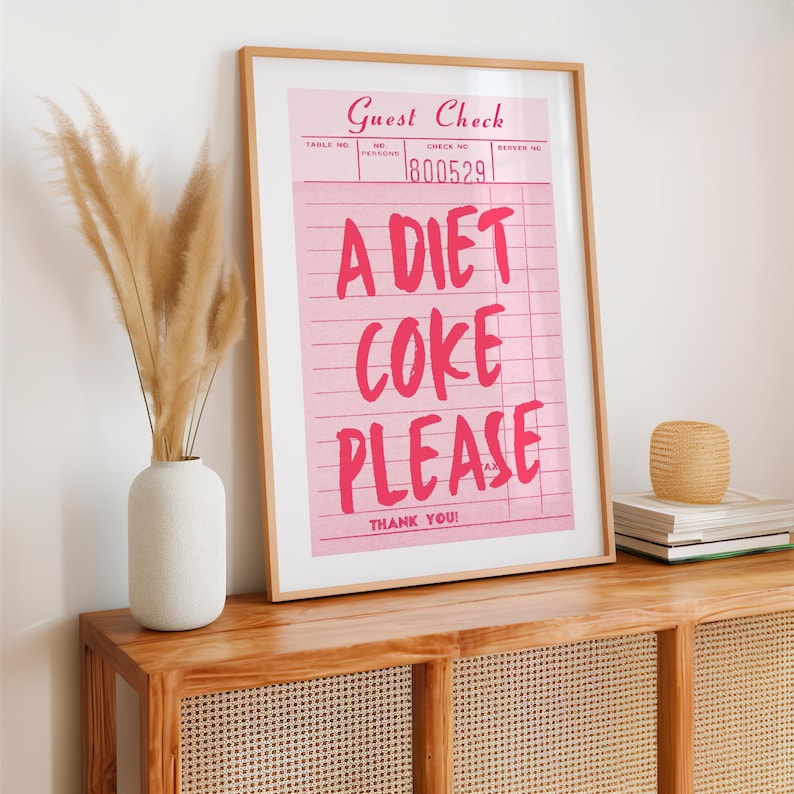 May include: A framed pink print resembling a guest check, with the words "A DIET COKE PLEASE" in bold pink letters. The print is displayed on a wooden cabinet, alongside a vase with dried plants.