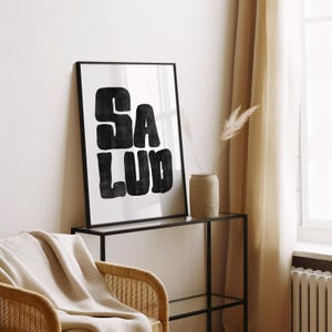 Op de afbeelding: Een zwart-witte ingelijste print met het woord "SALUD" in een vet, gestileerd lettertype. De print hangt aan een muur boven een zwart metalen plank.