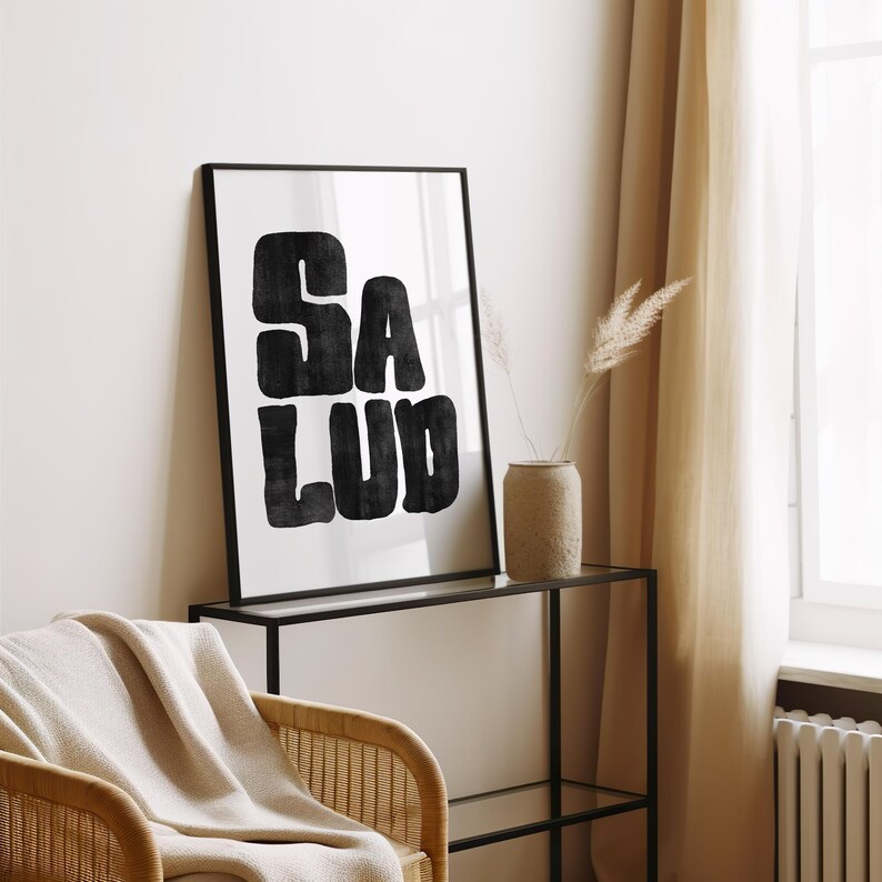 Op de afbeelding: Een zwart-witte ingelijste print met het woord "SALUD" in een vet, gestileerd lettertype. De print hangt aan een muur boven een zwart metalen plank.
