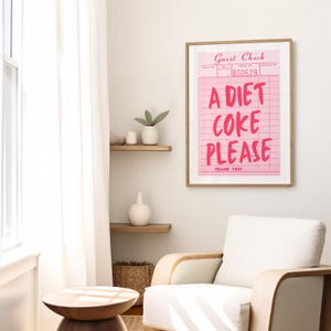 May include: A framed pink print with the words "A Diet Coke Please" in bold, bright pink letters. The print resembles a guest check and is displayed in a room with a white chair and wooden shelves.