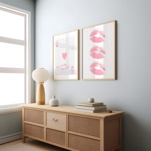 May include: Two framed art prints. One features a playing card with a heart and the text "How lucky are we!". The other print displays three pink lip imprints. Both are in light wood frames, displayed on a light wood cabinet.