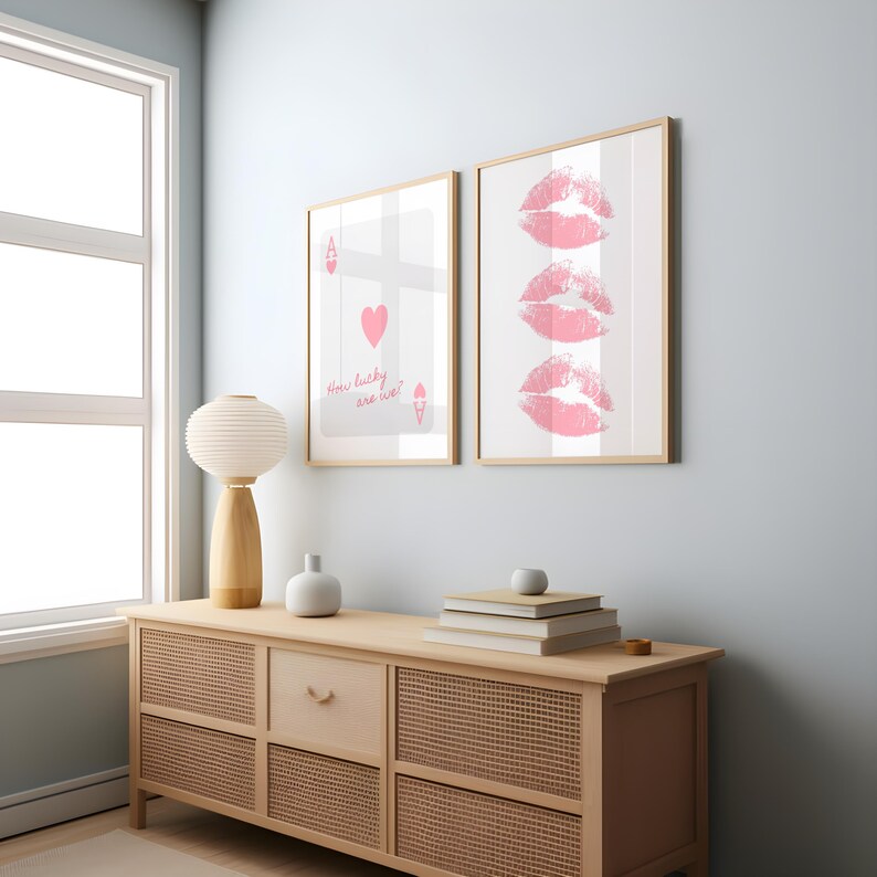 May include: Two framed art prints. One features a playing card with a heart and the text "How lucky are we!". The other print displays three pink lip imprints. Both are in light wood frames, displayed on a light wood cabinet.