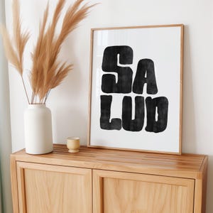 Op de afbeelding: Een ingelijst zwart-witdruk met het woord "SALUD" in vetgedrukte blokletters. De print hangt aan een licht houten muur boven een licht houten kast.